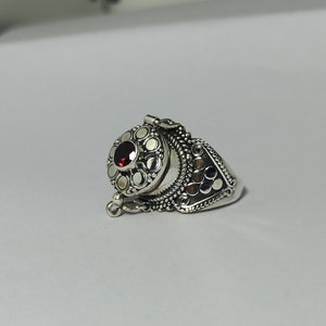 May include: A silver ring with a small, round, red gemstone set in the center of a circular design. The ring has a detailed, ornate design with a hinged lid.