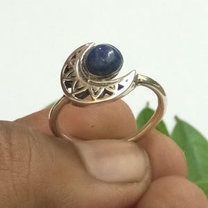 Könnte beinhalten: Ein silberfarbener Ring mit einem Mondsichel-Design, das komplizierte Ausschnitte aufweist und einen glatten, runden, tiefblauen Stein in der Mitte des Mondes trägt. Die Ringschiene ist dünn und poliert, was ein stimmiges Gesamtbild ergibt.