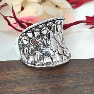 Könnte beinhalten: Ein silberner Ring mit einem strukturierten Design, das an Schuppen erinnert. Der Ring ist breit und hat eine gebogene Form.