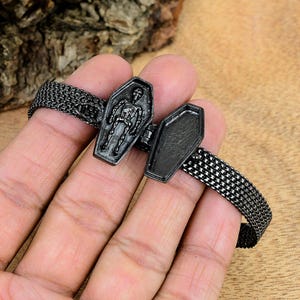 May include: A black bracelet featuring a coffin-shaped charm with a skeleton design. The charm opens to reveal an empty interior. The bracelet has a mesh chain design and is made of a dark metal.