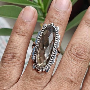 Puede incluir: Un anillo de plata con una gran piedra preciosa de cuarzo ahumado facetado de forma alargada. La piedra está engastada en un bisel de plata decorativo con un borde de cuentas, creando un marco texturizado alrededor del cristal marrón grisáceo.