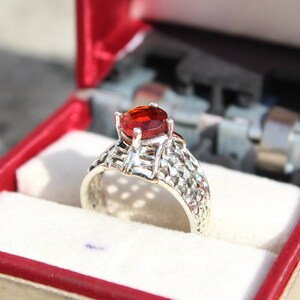 May include: A silver ring with a red gemstone set in a delicate, intricate design. The ring is displayed in a red velvet ring box.