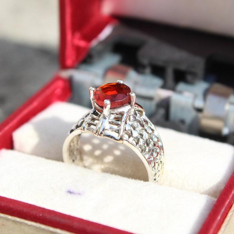 May include: A silver ring with a red gemstone set in a delicate, intricate design. The ring is displayed in a red velvet ring box.