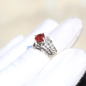 May include: A silver ring with a red gemstone set in a delicate, intricate design. The ring is on a white surface.