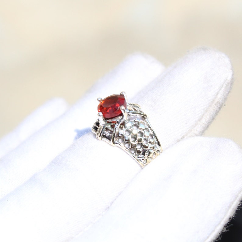 May include: A silver ring with a red gemstone set in a delicate, intricate design. The ring is on a white surface.