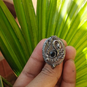 Puede incluir: Un anillo de plata con un diseño ovalado que presenta una representación detallada de un cisne con un ojo de piedra preciosa negra. El anillo está colocado sobre un fondo de hojas verdes.