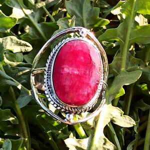 May include: A silver ring featuring a large, oval-shaped, vibrant pinkish-red gemstone set in a decorative, twisted metal border. The setting is surrounded by a smooth, circular silver frame that connects to the band, creating a layered, open design.