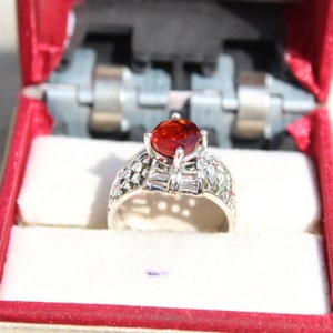 May include: A silver ring with a red gemstone set in a delicate, intricate design. The ring is displayed in a red velvet box.