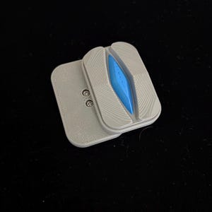 May include: A gray and white device with a blue center, possibly a phone accessory. The device has a rectangular shape with a textured surface and a small, attached gray piece. Two small silver screws are visible.