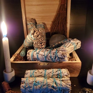 May include: A wooden box filled with bundles of white sage smudge sticks tied with blue string. The box is open and the sage sticks are spilling out. There are two candles and a small broom in the image.