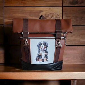 Könnte beinhalten: Ein brauner Lederrucksack mit einer Reißverschlusstasche vorne und einem weißen Patch mit einem Cartoon-Berner Sennenhund-Welpen. Der Rucksack hat zwei verstellbare Träger.