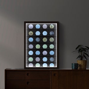 May include: A rectangular dark wood display case featuring six horizontal shelves illuminated by internal LED lighting. The case holds twenty-four circular coins or medallions arranged in four columns, showcasing various metallic tones and embossed designs.