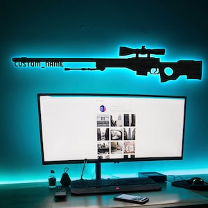 Puede incluir: Silueta negra de un rifle de francotirador de madera con el texto "CUSTOM_NAME" iluminado por una luz LED azul. El rifle está montado encima de un monitor de ordenador en un escritorio.