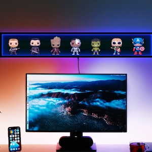 May include: A black wall-mounted shelf with blue LED lighting displays seven character figurines. Below, a computer monitor sits on a desk with a smartphone and a cup of tea. The wall is lit with a gradient of colors.
