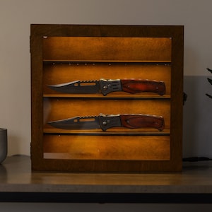 May include: A wooden display case with two folding knives with brown handles. The case has a glass front and is lit from within.