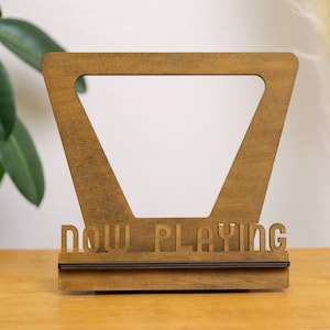May include: A wooden stand with a triangular cutout and the words "NOW PLAYING" on the front. The stand is brown and has a black trim.