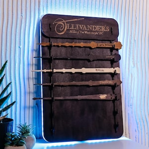 Könnte beinhalten: Ein schwarzer Holzständer mit dem Text "Ollivanders Makers of Fine Wands since 382 BC" und fünf Holzstäbe mit den Etiketten: "Expert Patronum", "Ashwinder", "Phoenixfeather", "Ebony/Garnet" und "Dragon Heartstring".
