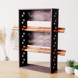 Pode incluir: Um suporte de madeira marrom escuro com um gráfico de punho branco e o texto "Hard Summer 2024" na frente. O suporte tem várias ranhuras para segurar baquetas de madeira.