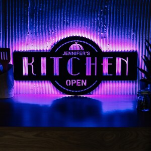 Könnte beinhalten: Schwarzes Küchenschild aus Metall mit dem Text "Jennifer's Kitchen Open", beleuchtet von violetten und blauen LED-Leuchten. Das Schild hat ein kreisförmiges Design mit einem stilisierten Bild eines Kochhutes.