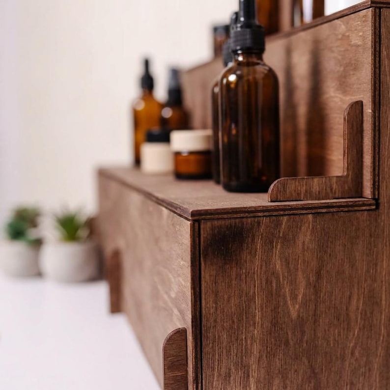 Peut inclure: Une &eacute;tag&egrave;re en bois brun fonc&eacute; &agrave; plusieurs niveaux, avec plusieurs flacons et pots en verre ambr&eacute;. L'&eacute;tag&egrave;re a une conception en gradins, chaque niveau contenant divers contenants. Le grain du bois est visible, et l'esth&eacute;tique g&eacute;n&eacute;rale est naturelle et minimaliste.