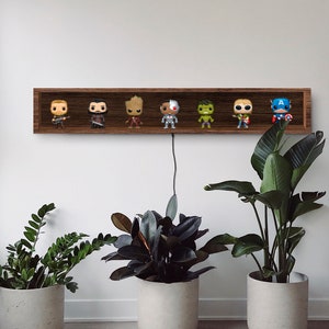 May include: A dark brown wooden shelf displays seven superhero Funko Pop figures. The shelf is mounted on a white wall, with a black cord hanging down. Potted plants in concrete planters are in the foreground.