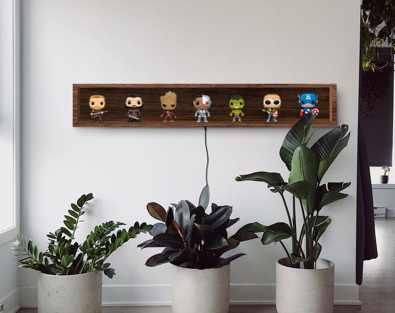 May include: A dark brown wooden shelf displays seven superhero Funko Pop figures. The shelf is mounted on a white wall, with a black cord hanging down. Potted plants in concrete planters are in the foreground.