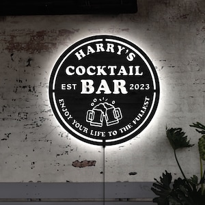 Puede incluir: Un letrero de pared circular negro con retroiluminación blanca que presenta el texto HARRY'S COCKTAIL BAR, EST 2023 y ENJOY YOUR LIFE TO THE FULLEST. El gráfico central muestra dos jarras de cerveza brindando con espuma en color blanco.