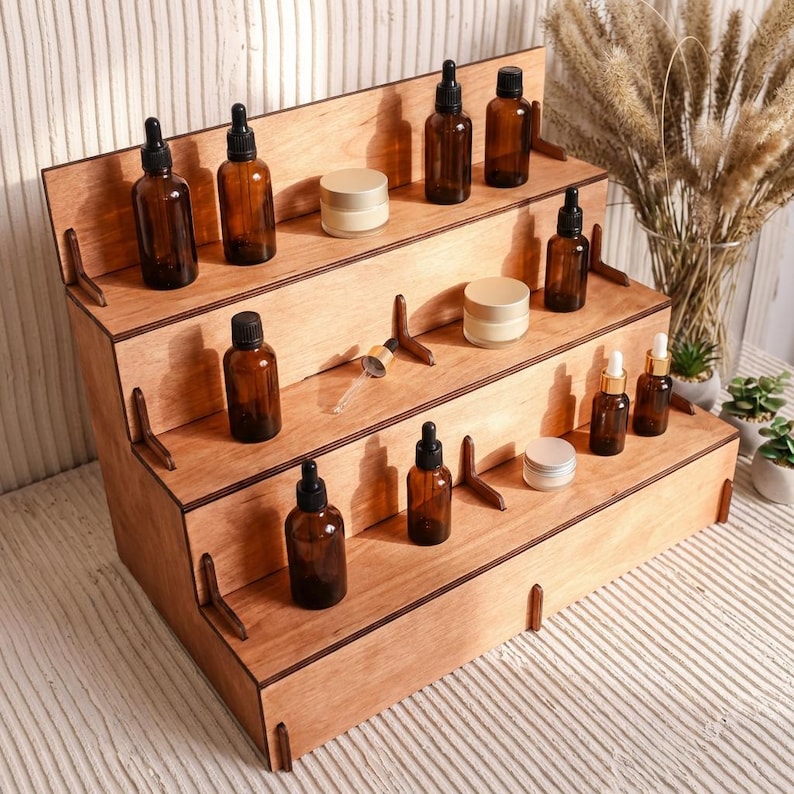 Peut inclure: Pr&eacute;sentoir en bois &agrave; plusieurs niveaux. Il pr&eacute;sente plusieurs &eacute;tag&egrave;res contenant des flacons en verre ambr&eacute; avec des compte-gouttes, de petits pots et des compte-gouttes en verre transparent. Le pr&eacute;sentoir est en bois clair.