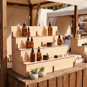 Könnte beinhalten: Ein mehrstufiger Holzständer mit braunen Glasflaschen mit Sprühköpfen und Tropfern sowie kleinen Gläsern. Die Präsentation steht auf einem Holztisch, davor kleine Topfpflanzen. Die Szene deutet auf eine natürliche Hautpflege- oder ätherische Öl-Produktpräsentation hin.