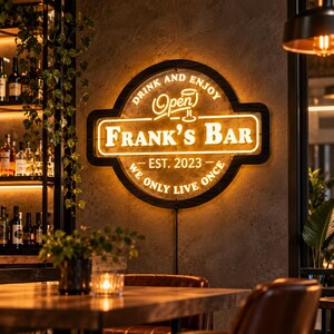 Op de afbeelding: Een cirkelvormig, verlicht wandbord met een zwarte rand en warm geel licht. De tekst luidt: DRINK AND ENJOY, Open, FRANK'S BAR, EST. 2023, WE ONLY LIVE ONCE. Het bord bevat een afbeelding van een glas boven de naam en hangt aan een muur.