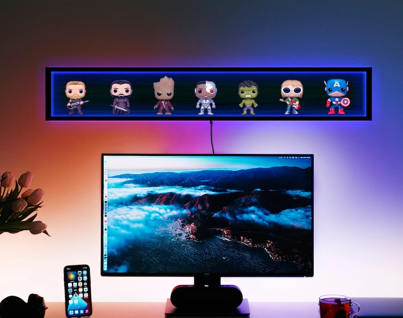 May include: A black wall-mounted shelf with blue LED lighting displays seven character figurines. Below, a computer monitor sits on a desk with a smartphone and a cup of tea. The wall is lit with a gradient of colors.