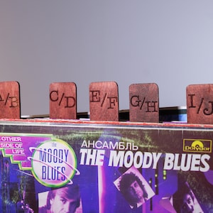 Puede incluir: Separadores de discos de vinilo de madera con las letras A/B, C/D, E/F, G/H e I/J, colocados sobre una pila de discos. La portada del álbum muestra "The Moody Blues" y "The Other Side of Life".