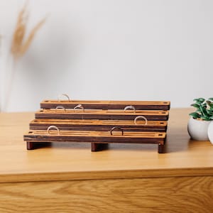May include: A wooden ring display stand with multiple tiers. The stand is made of dark wood with a natural wood finish. There are several rings on the stand, including silver and gold bands.