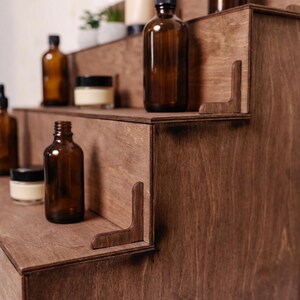 Peut inclure: Pr&eacute;sentoir en bois &agrave; plusieurs niveaux, de couleur marron fonc&eacute;. Il pr&eacute;sente des flacons en verre ambr&eacute; et de petits pots avec des couvercles noirs sur chaque niveau. Des supports d'angle d&eacute;coratifs compl&egrave;tent le design.