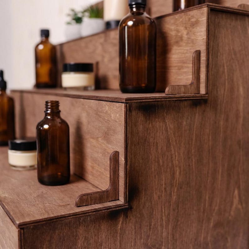 Peut inclure: Pr&eacute;sentoir en bois &agrave; plusieurs niveaux, de couleur marron fonc&eacute;. Il pr&eacute;sente des flacons en verre ambr&eacute; et de petits pots avec des couvercles noirs sur chaque niveau. Des supports d'angle d&eacute;coratifs compl&egrave;tent le design.