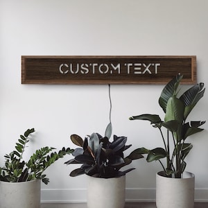 May include: A rectangular wooden sign with the words "CUSTOM TEXT" in white, mounted on a white wall. The sign is illuminated and has a power cord. Three potted plants are in front of the wall.