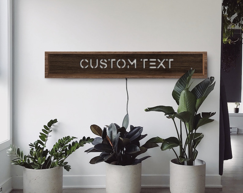 May include: A rectangular wooden sign with the words "CUSTOM TEXT" in white, mounted on a white wall. The sign is illuminated and has a power cord. Three potted plants are in front of the wall.