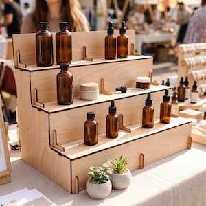 Peut inclure: Pr&eacute;sentoir en bois &agrave; plusieurs niveaux avec des flacons en verre ambr&eacute;, des pipettes et de petits pots. Le pr&eacute;sentoir est sur une table, avec de petites plantes en pot devant. Le tout est install&eacute; sur un march&eacute; en plein air.