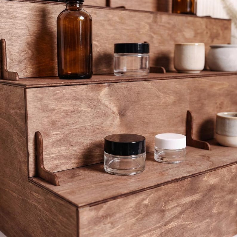Peut inclure: Pr&eacute;sentoir en bois &agrave; plusieurs niveaux avec divers petits pots en verre et r&eacute;cipients en c&eacute;ramique. Les pots ont des couvercles noirs et blancs. Une bouteille en verre brun est &eacute;galement visible. Le bois est teint&eacute; brun-rouge&acirc;tre chaud.