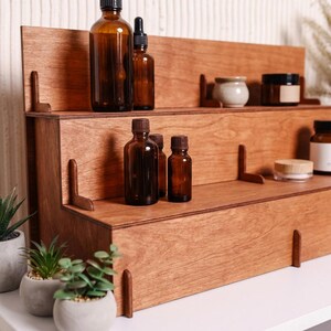 Peut inclure: Une &eacute;tag&egrave;re en bois &agrave; trois niveaux avec une finition en bois naturel. Les &eacute;tag&egrave;res contiennent diverses bouteilles, pots et petits pots en verre ambr&eacute;. L'&eacute;tag&egrave;re est con&ccedil;ue pour pr&eacute;senter des huiles essentielles, des cosm&eacute;tiques ou d'autres petits articles. L'esth&eacute;tique g&eacute;n&eacute;rale est minimaliste et fonctionnelle.