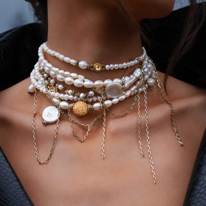 Può includere: Una collana di perle a strati con accenti dorati e dettagli a catena. La collana presenta una varietà di dimensioni e forme di perle, tra cui un grande ciondolo di perla piatta.