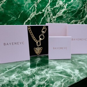 May include: Three white boxes with the text "BAYEMEYC" printed on them. The middle box contains a gold chain necklace with a leopard head pendant.