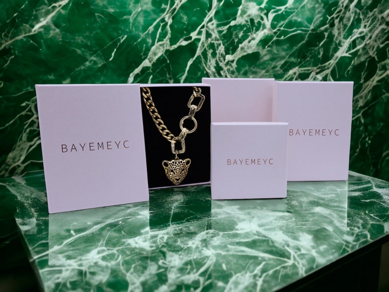 May include: Three white boxes with the text "BAYEMEYC" printed on them. The middle box contains a gold chain necklace with a leopard head pendant.