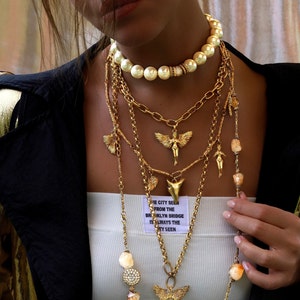May include: A woman wearing a white tank top with a gold chain necklace with a variety of charms, including a pearl necklace, a winged angel pendant, and a shark tooth pendant. The text on the tank top reads "The city seen from the Brooklyn Bridge is always the city seen."