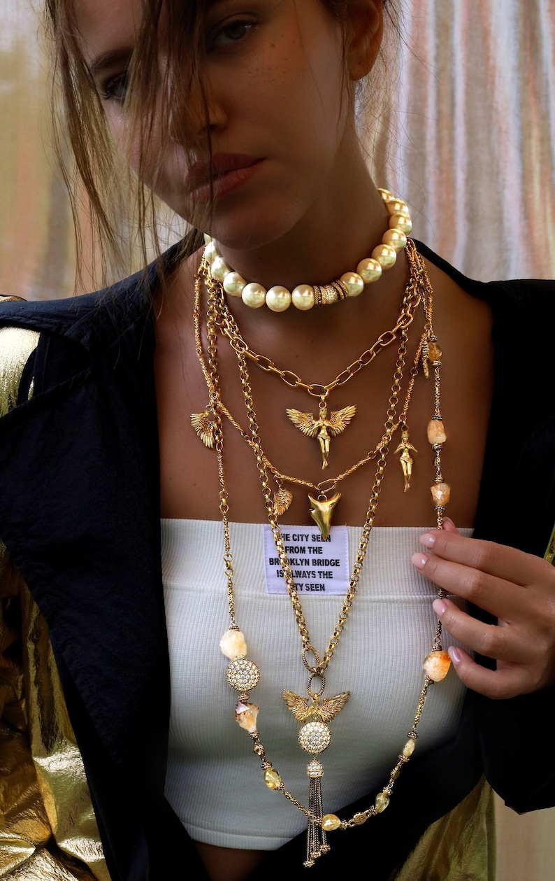 May include: A woman wearing a white tank top with a gold chain necklace with a variety of charms, including a pearl necklace, a winged angel pendant, and a shark tooth pendant. The text on the tank top reads "The city seen from the Brooklyn Bridge is always the city seen."