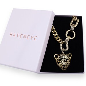 May include: A gold chain necklace with a large, detailed, gold jaguar head pendant. The necklace is in a white box with the word "BAYEMEYC" printed on the lid.