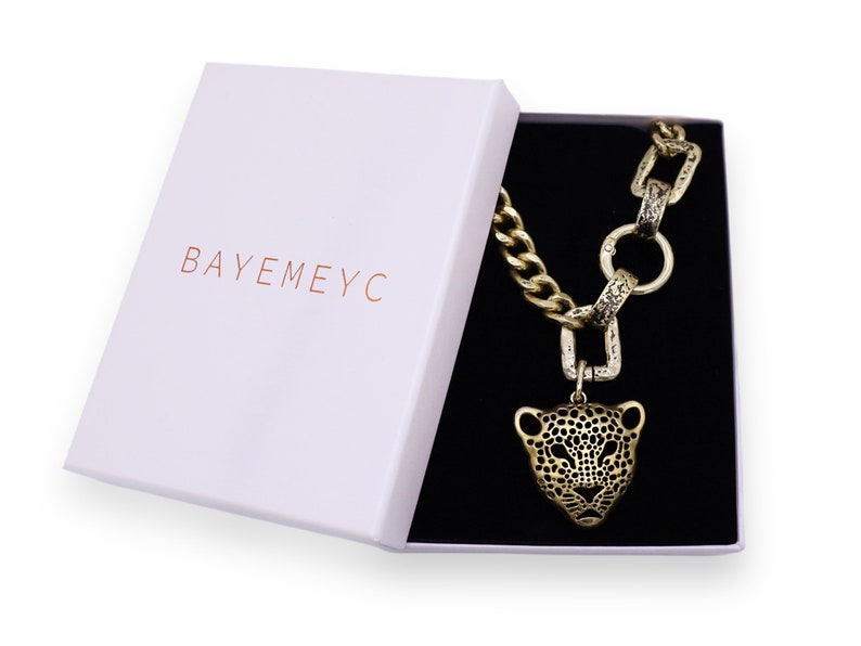 May include: A gold chain necklace with a large, detailed, gold jaguar head pendant. The necklace is in a white box with the word "BAYEMEYC" printed on the lid.