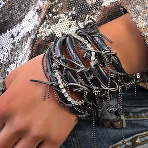Pode incluir: Uma pulseira de couro azul escuro com detalhes em metal prateado, correntes e strass. A pulseira é enrolada com arame prateado e apresenta pingentes de contas penduradas. O fundo inclui um top com lantejoulas e jeans.