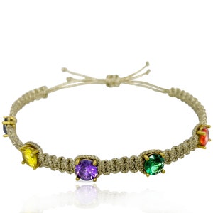May include: A beige braided bracelet with five colorful gemstones. The gemstones are purple, yellow, green, and red.