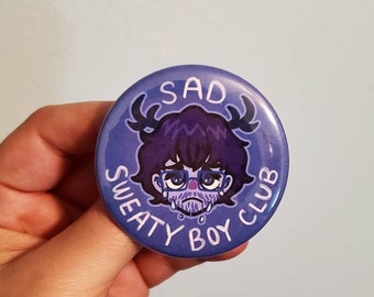 May include: A purple button with a cartoon illustration of a sad character with antlers, glasses, and a beard. The text "SAD SWEATY BOY CLUB" surrounds the character.